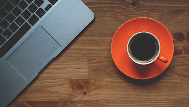 Cup of coffee on saucer beside MacBook on wooden table