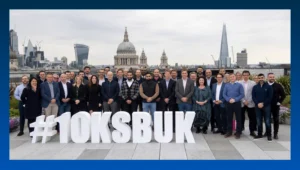 A diverse group of professionals posing on a rooftop in London with large 3D white letters in the foreground spelling out "#10KSBUK." The London skyline, including St. Paul's Cathedral and the Shard, is visible in the background under an overcast sky.